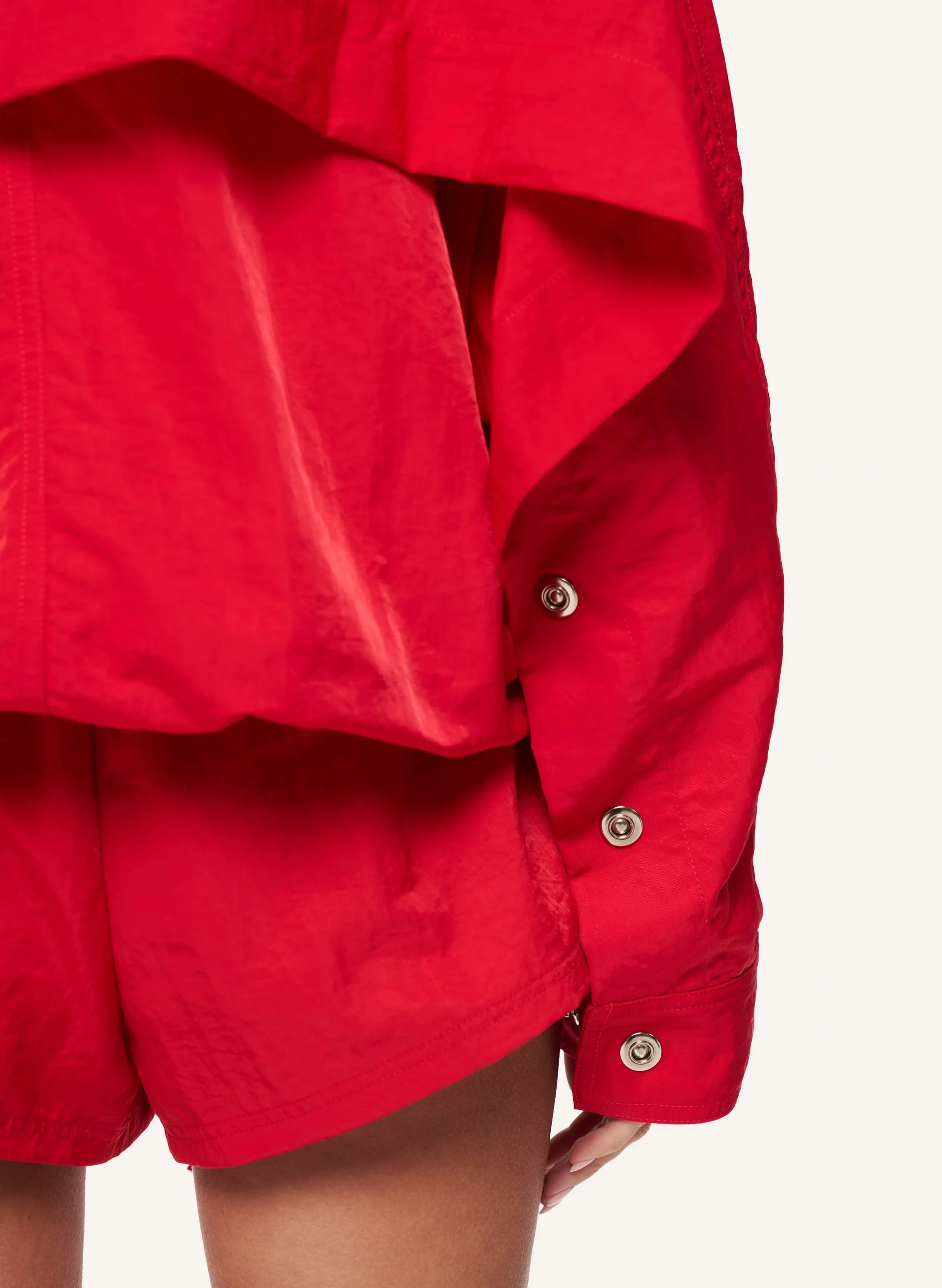 Close-up of a bright red matching set featuring a draped long-sleeve top with decorative buttons and matching shorts, shown on a model against a light off-white background.