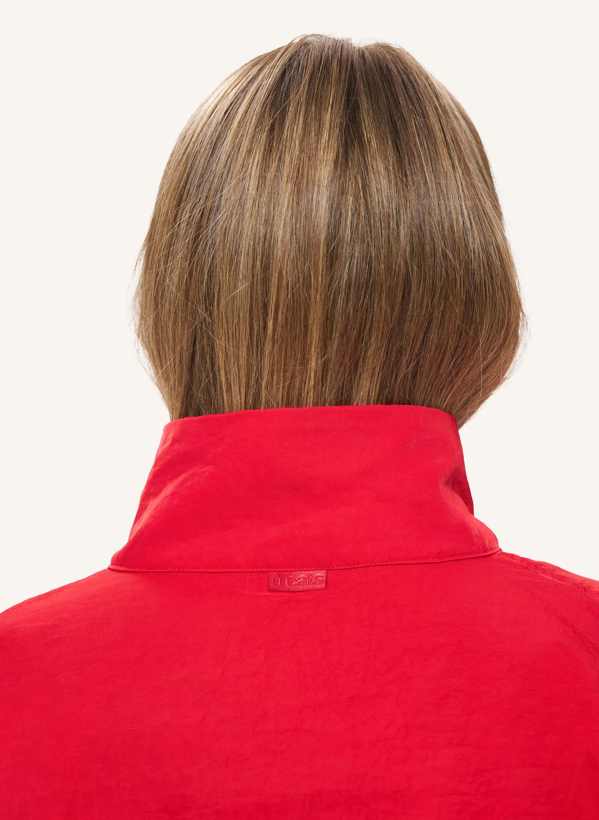 Back view of a person with shoulder-length brown hair wearing a bright red high-neck top against a plain light background.