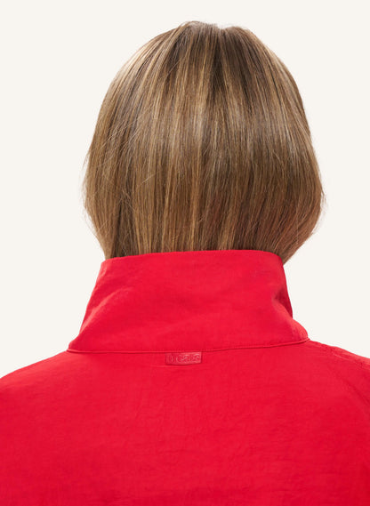 Back view of a person with shoulder-length brown hair wearing a bright red high-neck top against a plain light background.