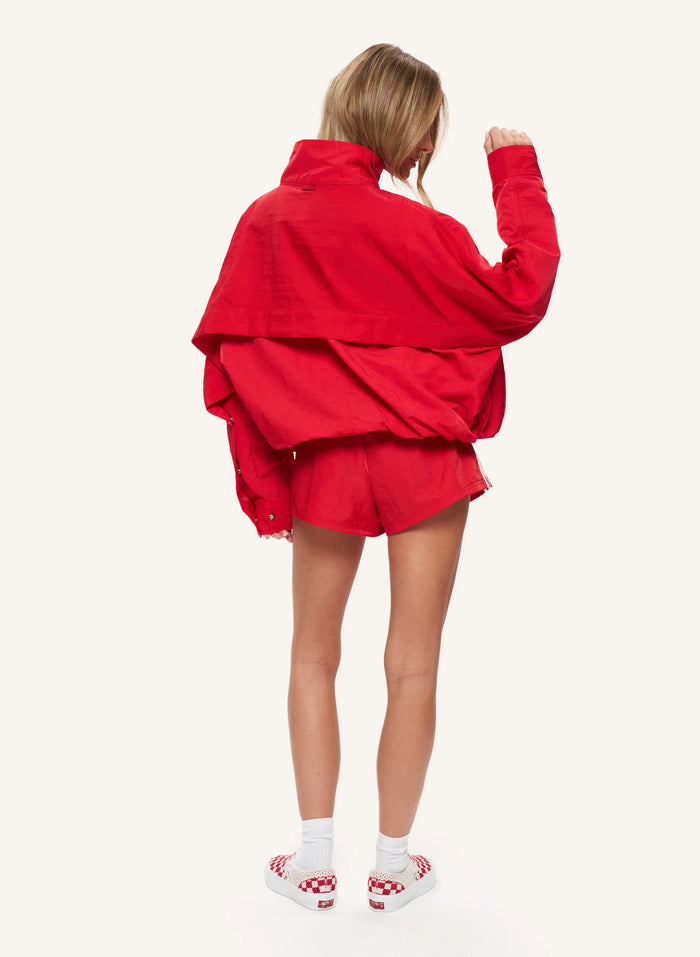 A model shown from behind wearing a bright red oversized jacket and matching red shorts, paired with white socks and red-and-white checkered slip-on shoes, against a plain off-white studio background.