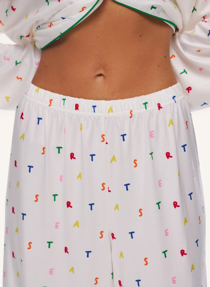 White pajama pants with an elastic waistband and a colorful repeating pattern of letters from the Stranger Things series, shown on a model.