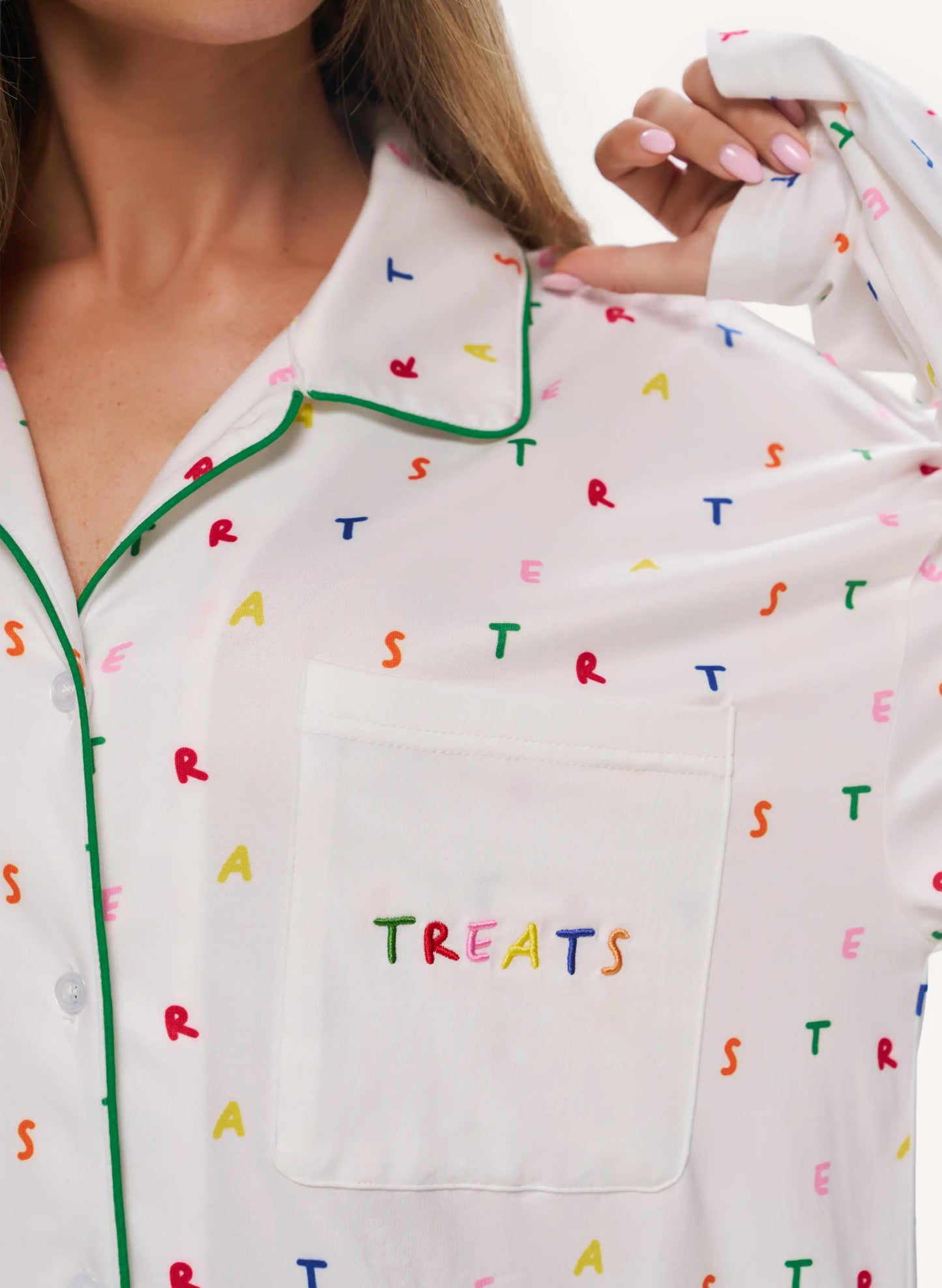 White pajama top with a colorful alphabet print and green piping, featuring a chest pocket with 'TREATS' embroidered in multicolored letters. A model is shown wearing and holding the pajama shirt.