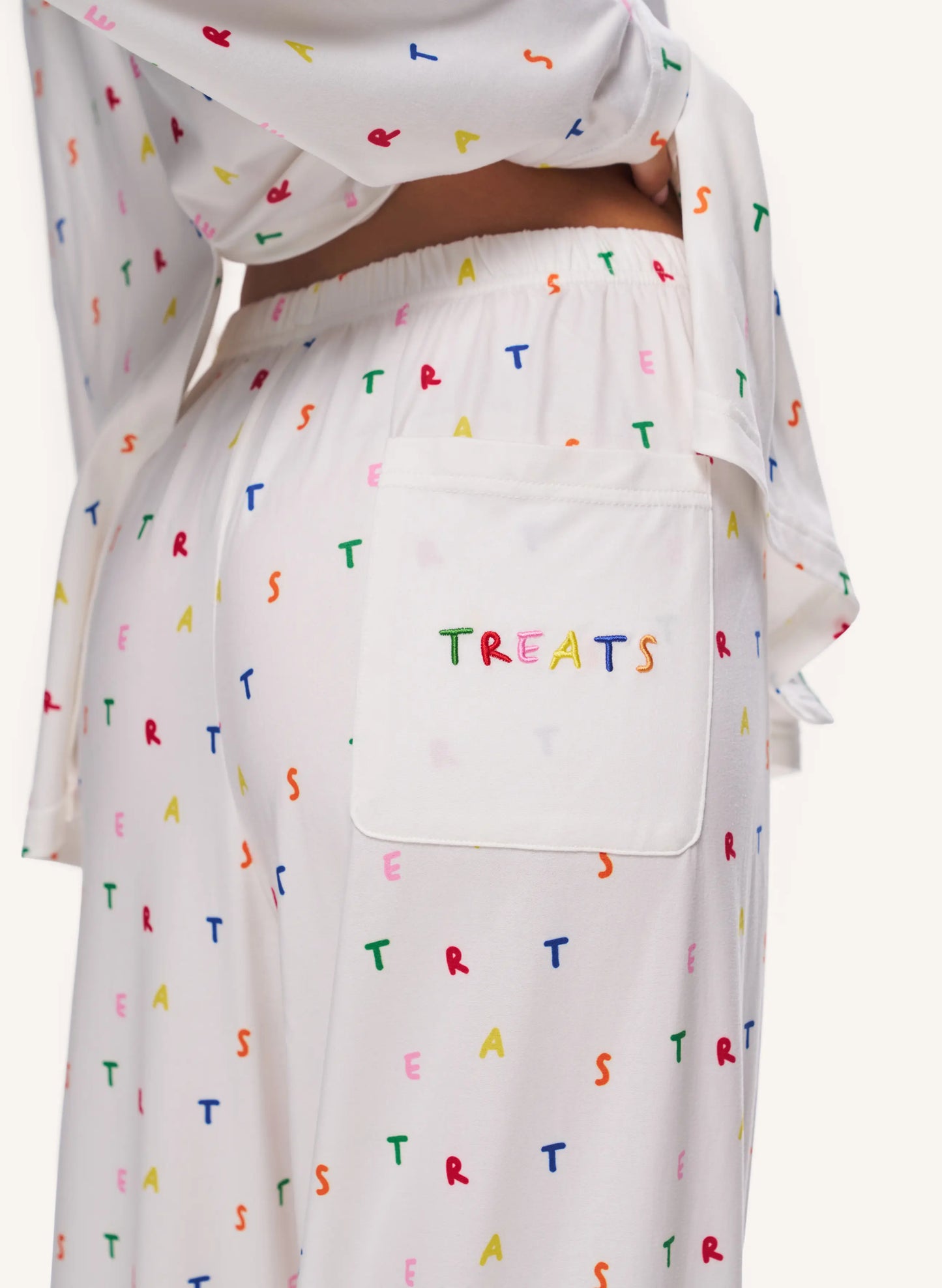 White sleepwear set with a colorful alphabet 'TREATS' print, featuring a large patch pocket on the pants with 'TREATS' embroidered in multicolored letters.