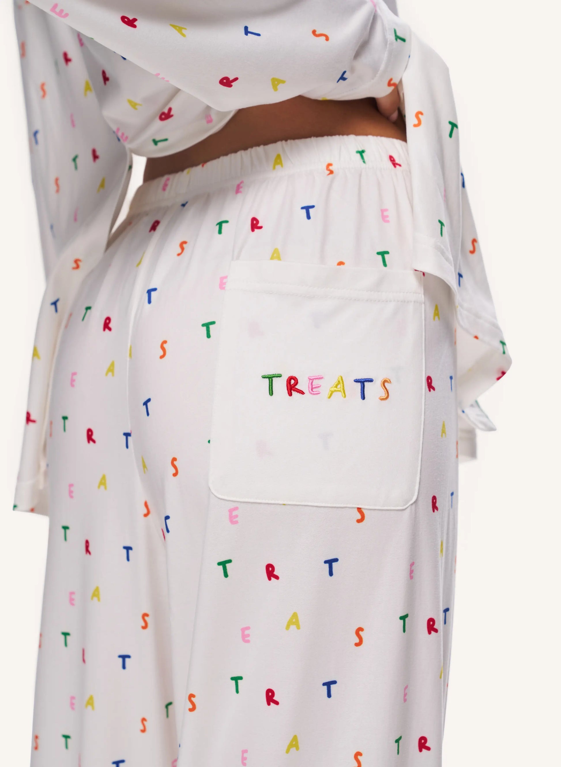 White sleepwear set with a colorful alphabet 'TREATS' print, featuring a large patch pocket on the pants with 'TREATS' embroidered in multicolored letters.