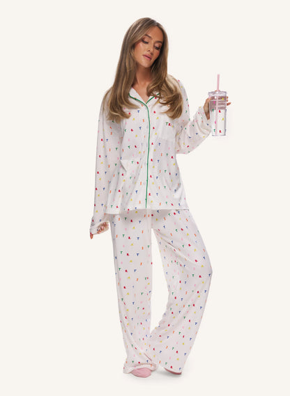 A woman modeling a two-piece white pajama set with a colorful confetti print, holding a clear tumbler with a pink straw against a solid off-white background.