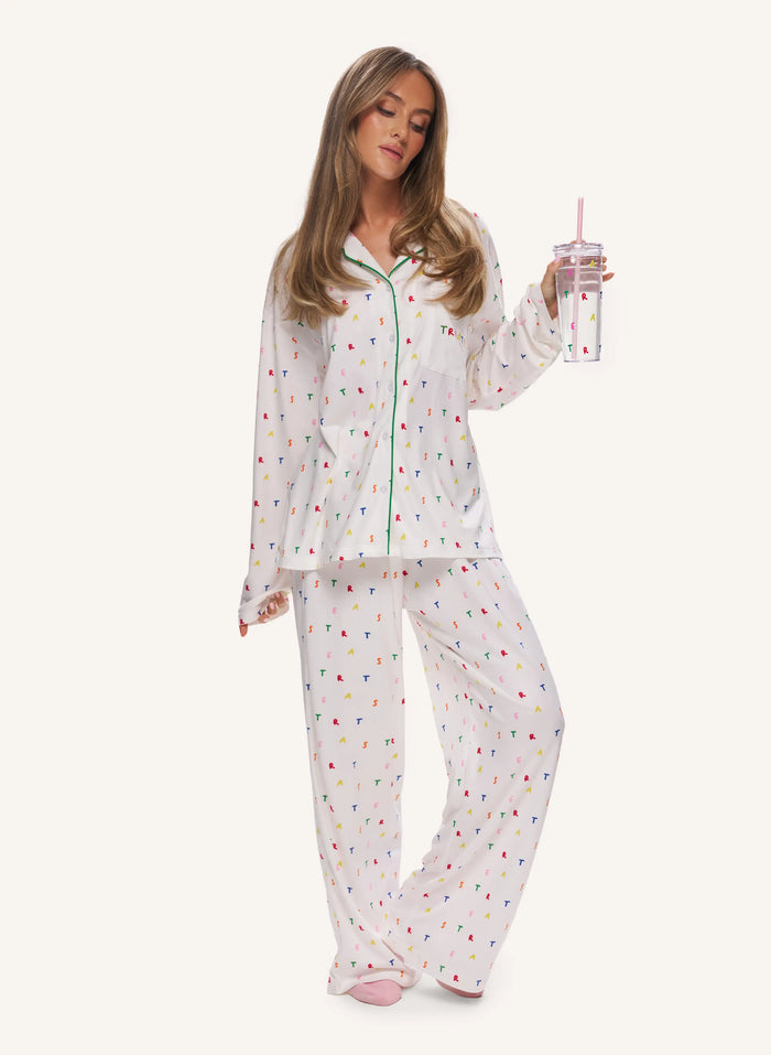 A woman modeling a two-piece white pajama set with a colorful confetti print, holding a clear tumbler with a pink straw against a solid off-white background.