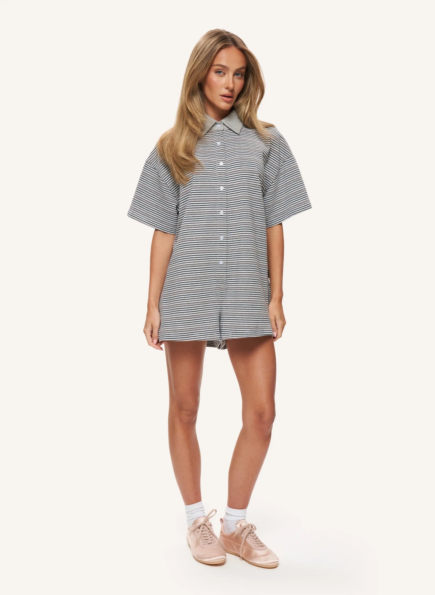 A woman modeling a short-sleeve button-front shirt dress with thin gray and white horizontal stripes, paired with white socks and light pink sneakers, against a solid off-white background.
