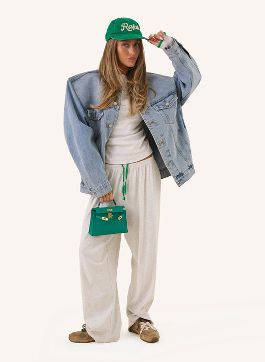 A woman modeling a light blue oversized denim jacket, cream sweatshirt, and cream wide-leg sweatpants, styled with a green baseball cap, a small green handbag, and brown suede shoes against a solid off-white background.