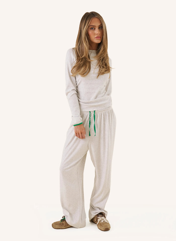 A woman modeling a matching two-piece loungewear set consisting of a light beige long-sleeve knit top and wide-leg sweatpants with green drawstrings, paired with brown suede shoes, against a solid off-white background.