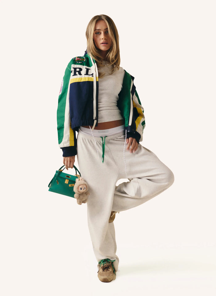 A model poses in a studio setting wearing a green, navy, and yellow color-blocked bomber jacket with 'RL' lettering on the chest, paired with cream sweatpants and holding a green handbag.