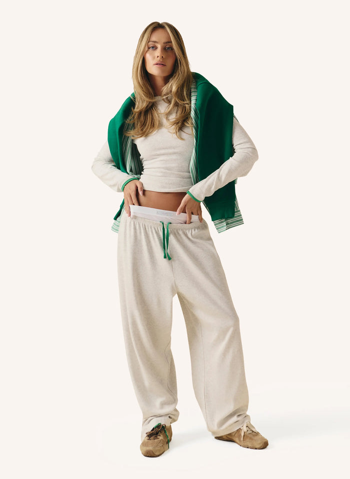 A woman modeling a white ribbed long-sleeve crop top, cream sweatpants with a green drawstring, and a dark green cardigan draped over her shoulders, set against a solid off-white background.