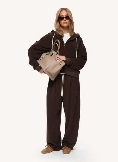 A model wearing a dark brown zip-up hoodie and matching sweatpants, holding a beige handbag, photographed against a solid off-white background.