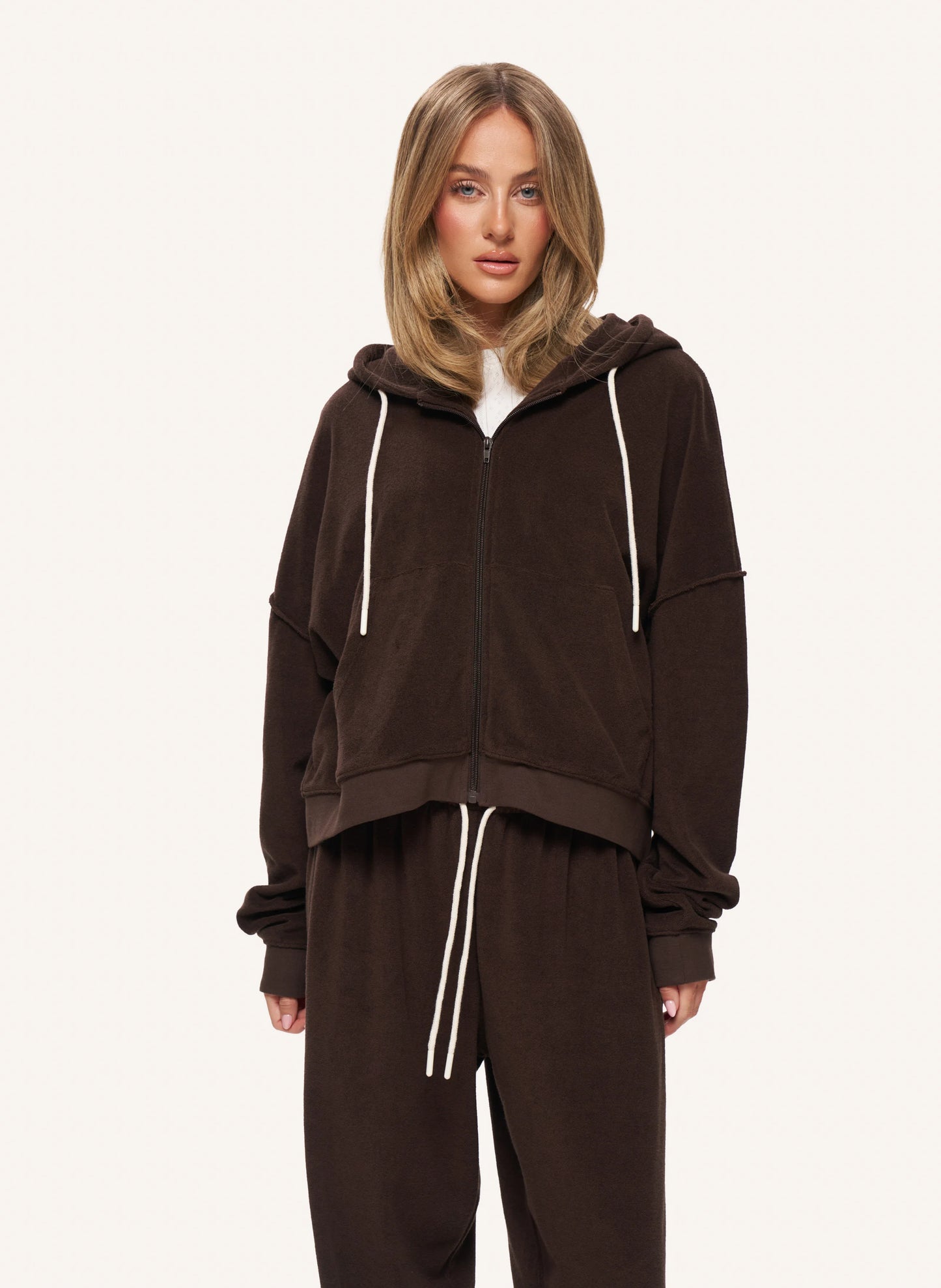 A woman modeling a dark brown zip-up hoodie and matching sweatpants set with white drawstrings, photographed against a clean off-white studio background.