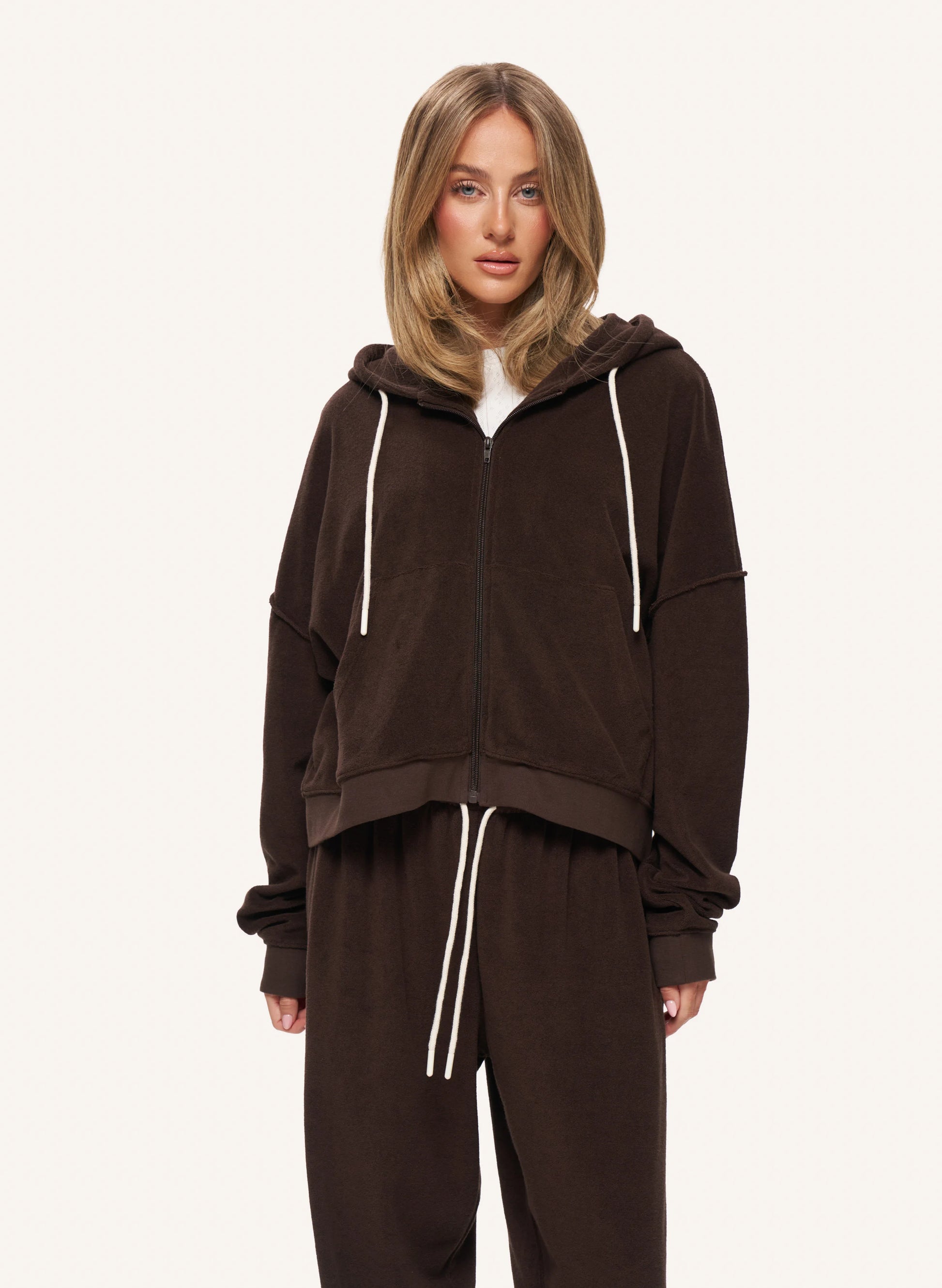 A woman modeling a dark brown zip-up hoodie and matching sweatpants set with white drawstrings, photographed against a clean off-white studio background.