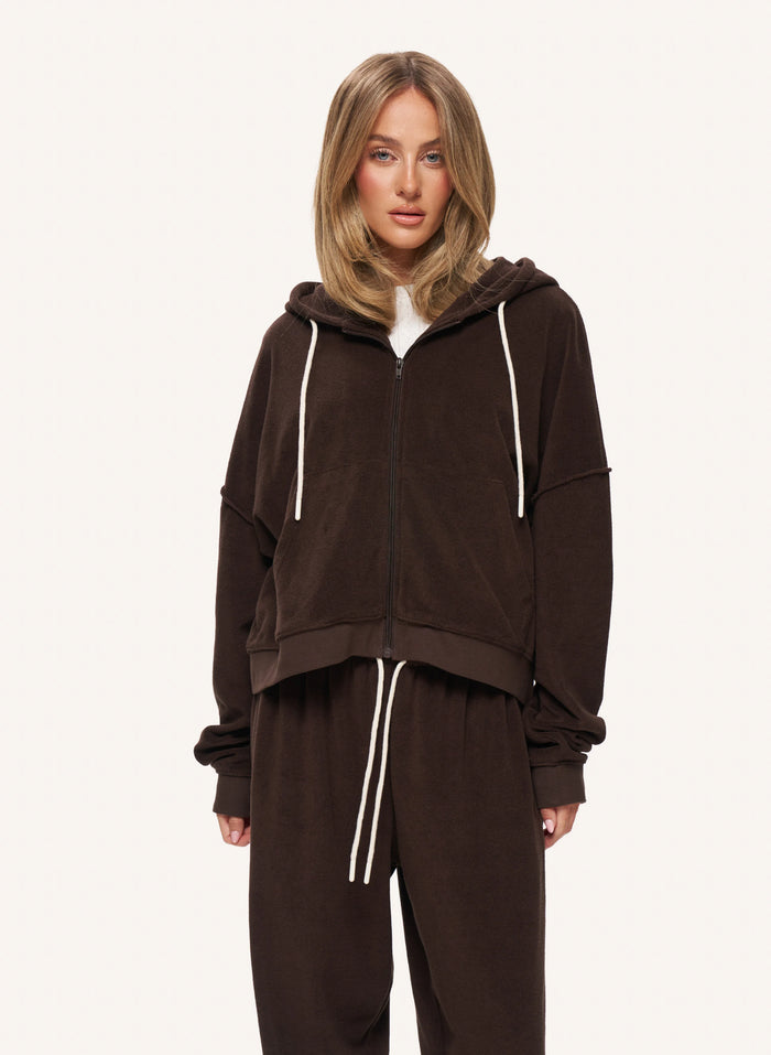 A woman modeling a dark brown zip-up hoodie and matching sweatpants set with white drawstrings, photographed against a clean off-white studio background.