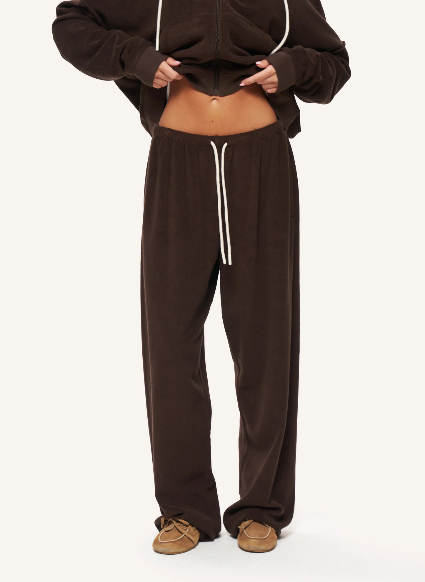A model wearing dark brown lounge pants with white drawstrings and a matching dark brown cropped hoodie, paired with tan slip-on shoes, against a plain off-white background.