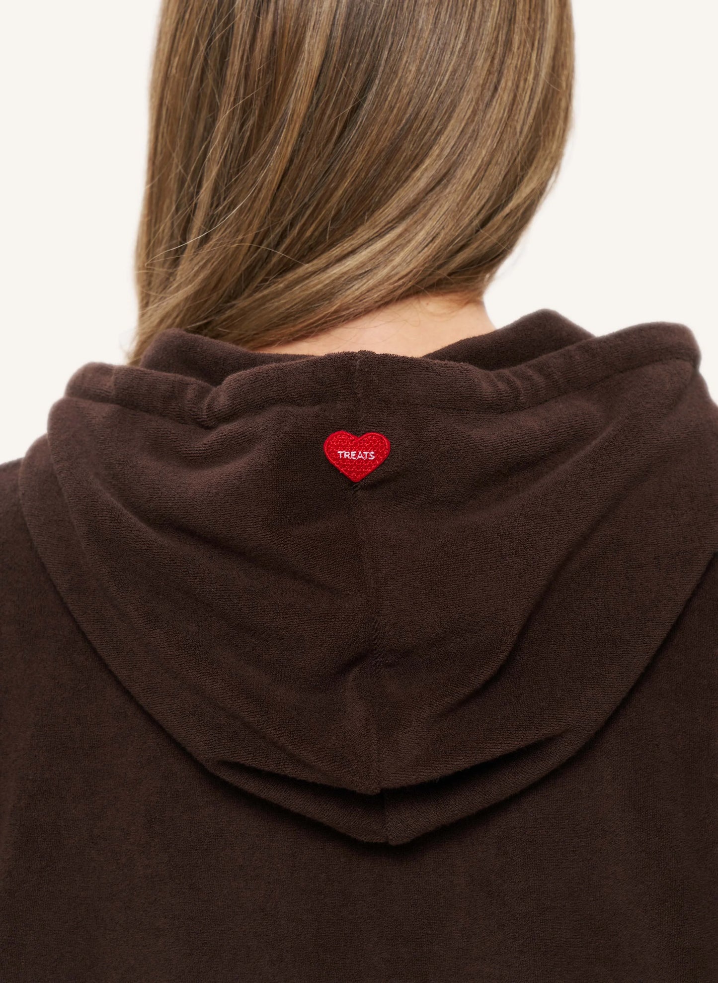 Close-up back view of a person wearing a dark brown hooded sweatshirt, showing a small red heart-shaped patch with the word 'TREATS' on the back of the neck.