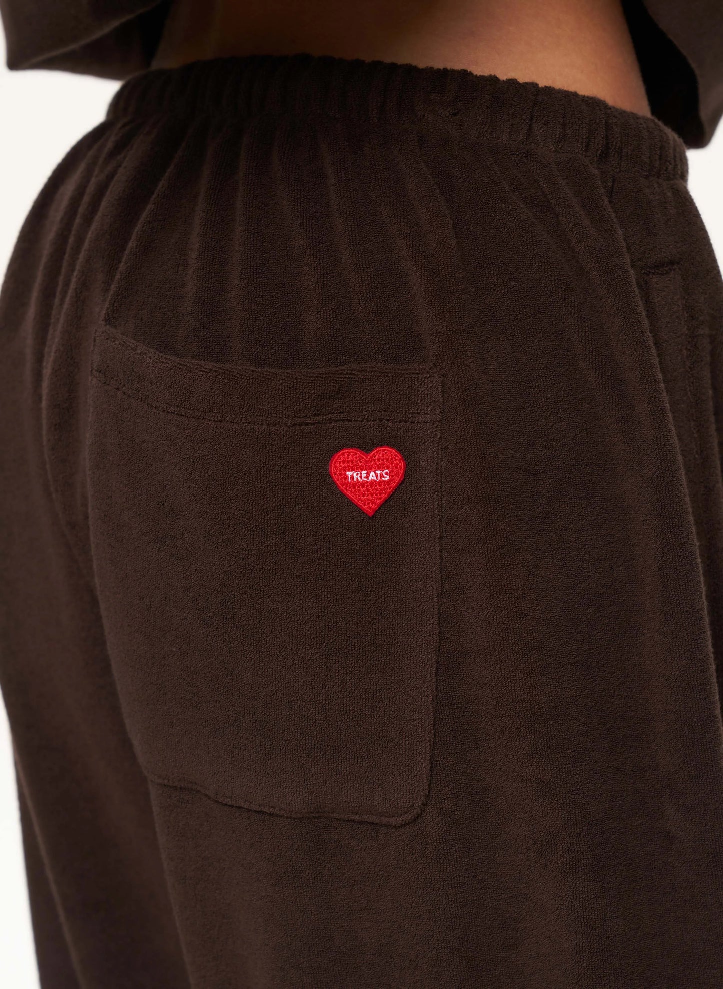 Close-up of dark brown corduroy sweatpants featuring a side pocket with a red embroidered heart containing the white text 'TREATS'.