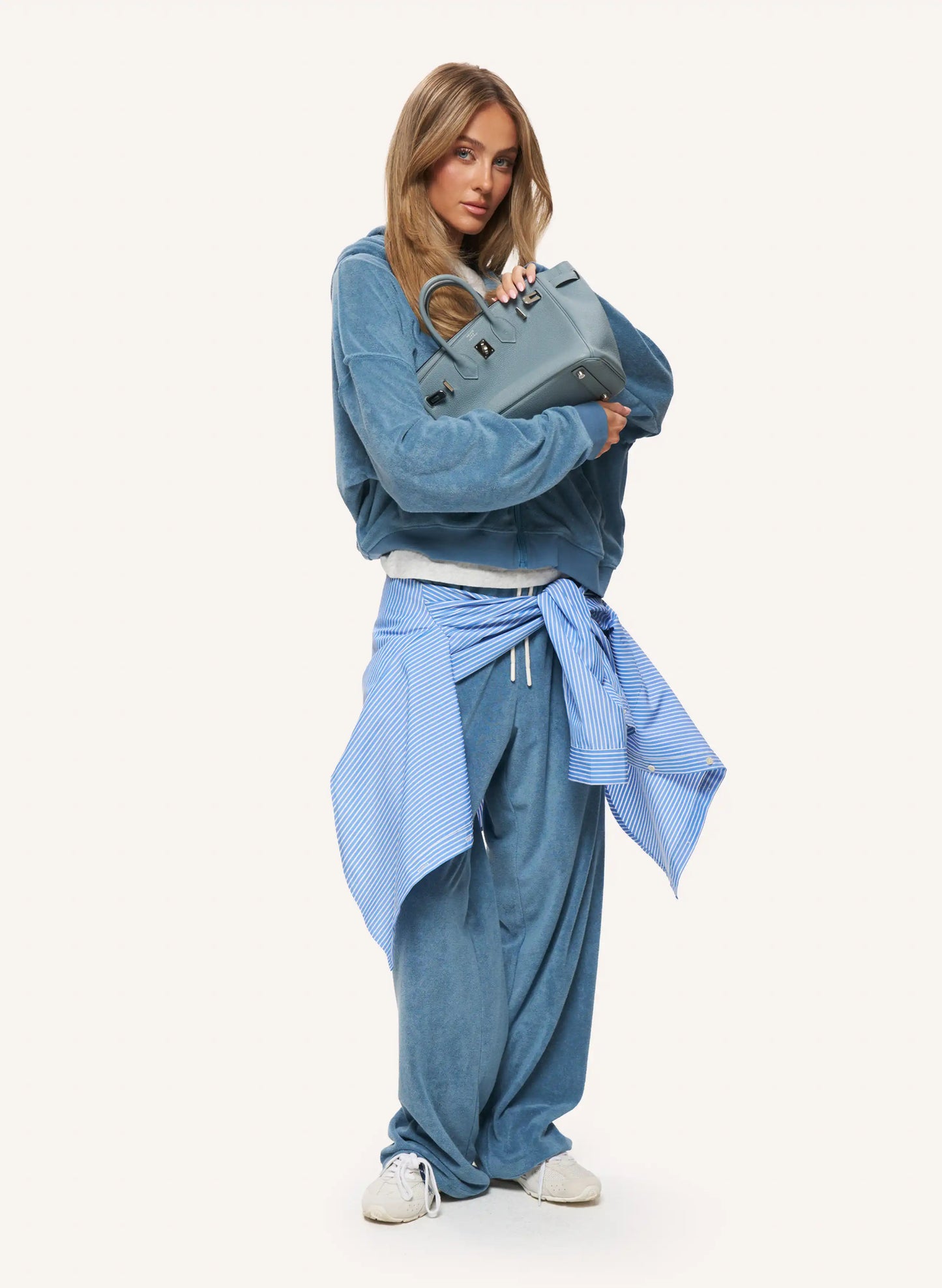 A model stands against a plain light background wearing a blue denim long-sleeve top, matching wide-leg jeans, a light blue striped shirt tied around her waist, and white sneakers, while holding a blue-gray handbag.