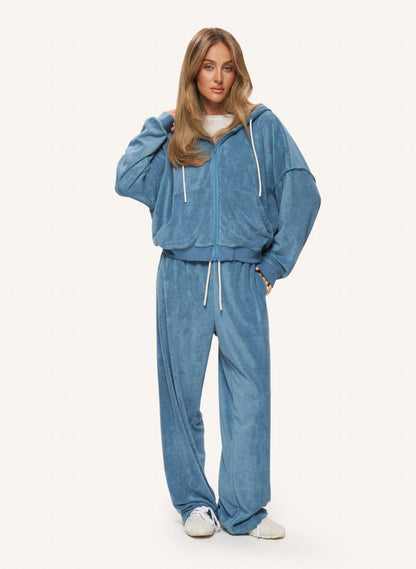 A woman modeling a matching blue velour lounge set consisting of a zip-up hoodie and wide-leg sweatpants, styled with white sneakers against a plain off-white background.