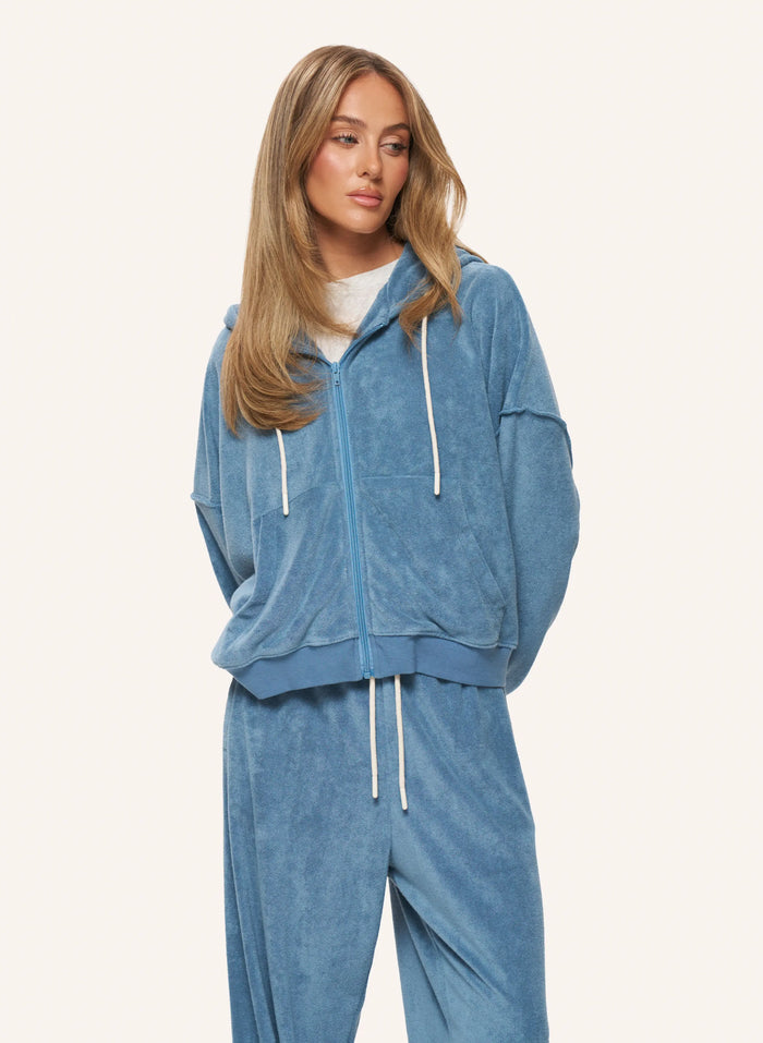 A woman modeling a matching blue velour lounge set consisting of a zip-up hoodie and sweatpants, styled with white drawstrings over a white t-shirt, against a solid off-white background.