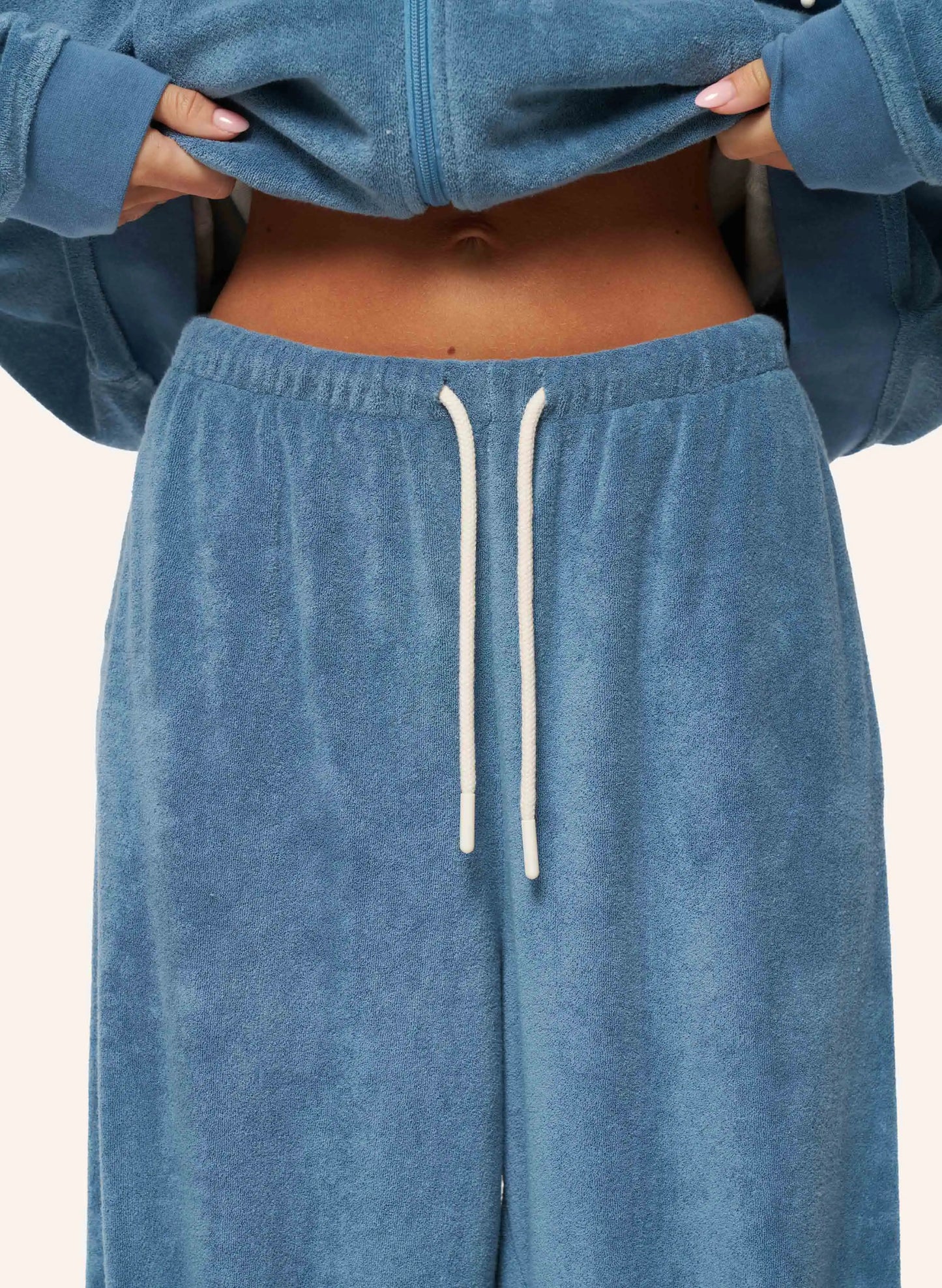 A close-up of a person wearing a matching dusty blue velour lounge set, featuring a cropped zip-up hoodie and wide-leg sweatpants with a white drawstring.
