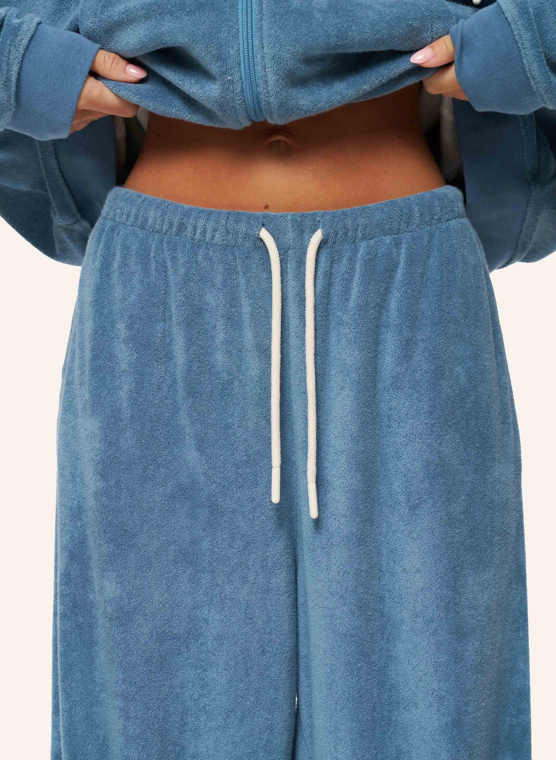 A close-up of a person wearing a matching dusty blue velour lounge set, featuring a cropped zip-up hoodie and wide-leg sweatpants with a white drawstring.
