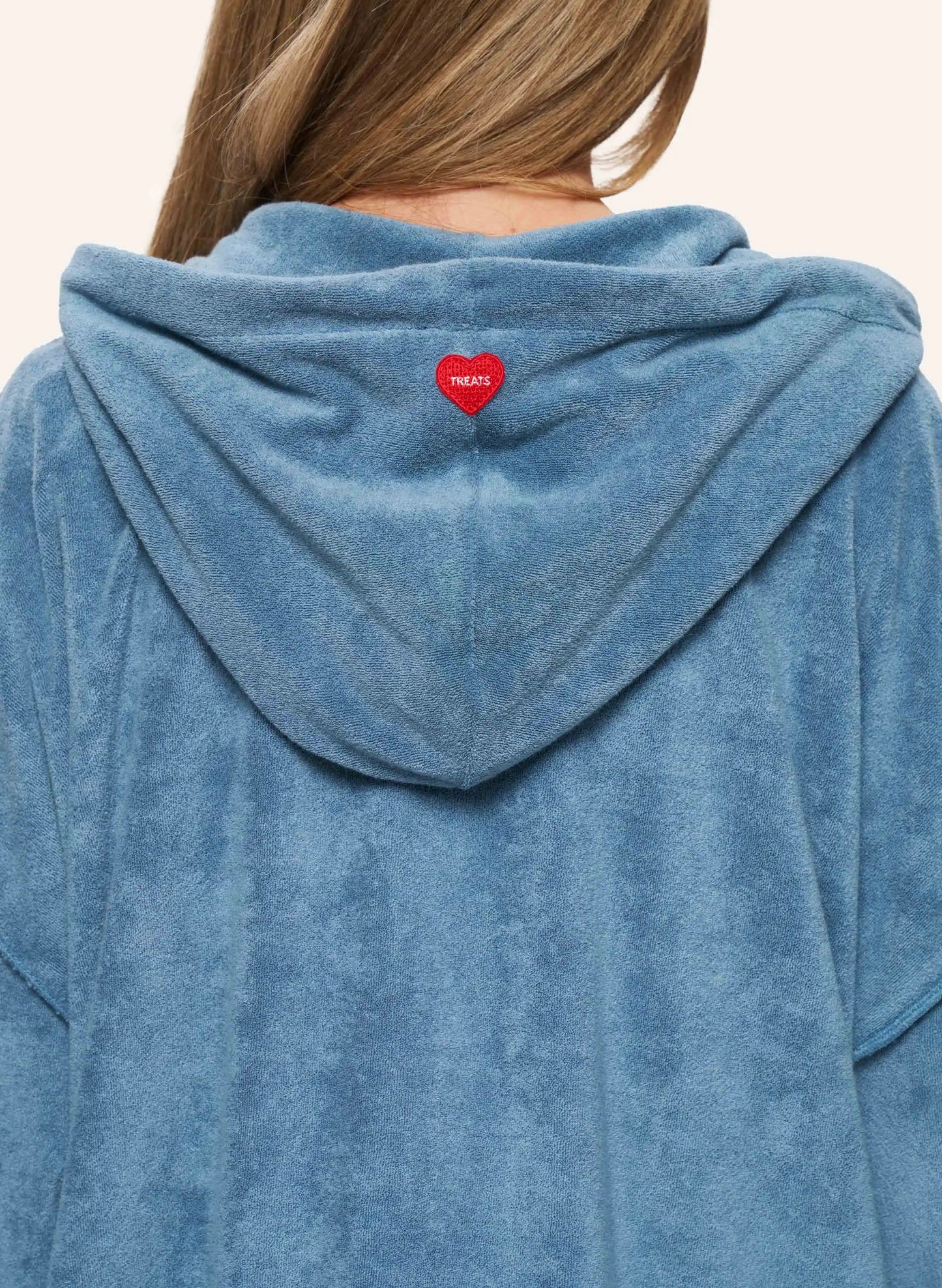 A close-up back view of a person wearing a dusty blue plush hooded sweatshirt. A small red heart-shaped patch with white text is centered on the back of the hood.