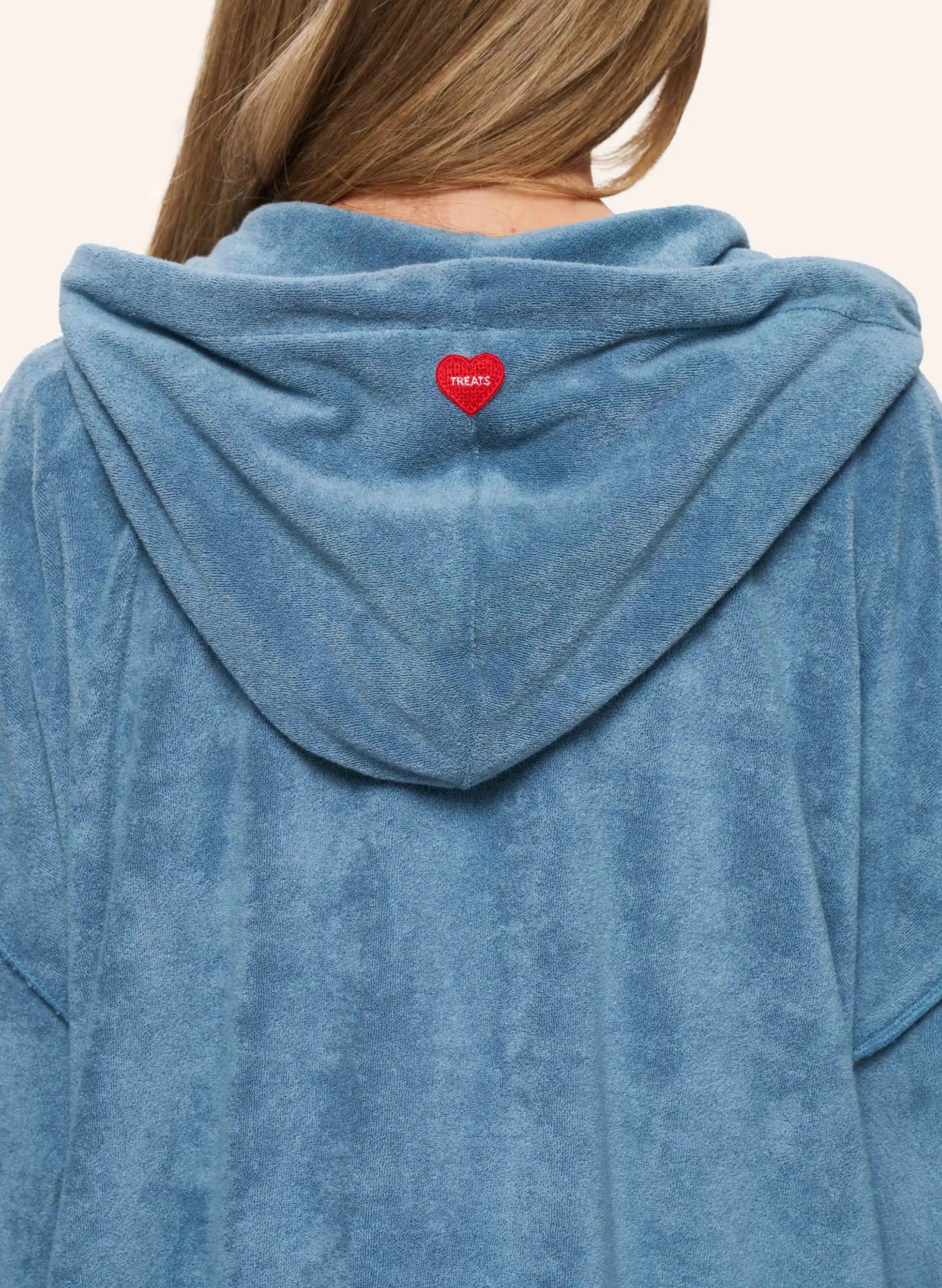 A close-up back view of a person wearing a dusty blue plush hooded sweatshirt. A small red heart-shaped patch with white text is centered on the back of the hood.
