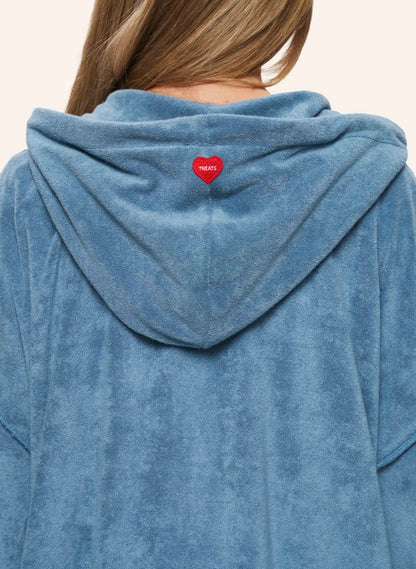 A close-up back view of a person wearing a dusty blue plush hooded sweatshirt. A small red heart-shaped patch with white text is centered on the back of the hood.