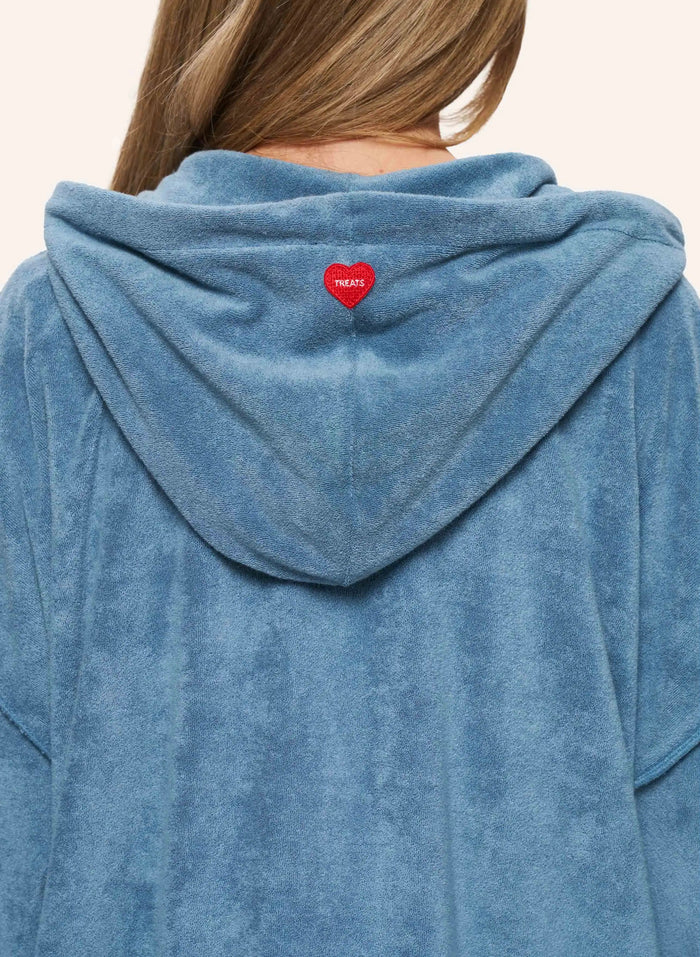 A close-up back view of a person wearing a dusty blue plush hooded sweatshirt. A small red heart-shaped patch with white text is centered on the back of the hood.