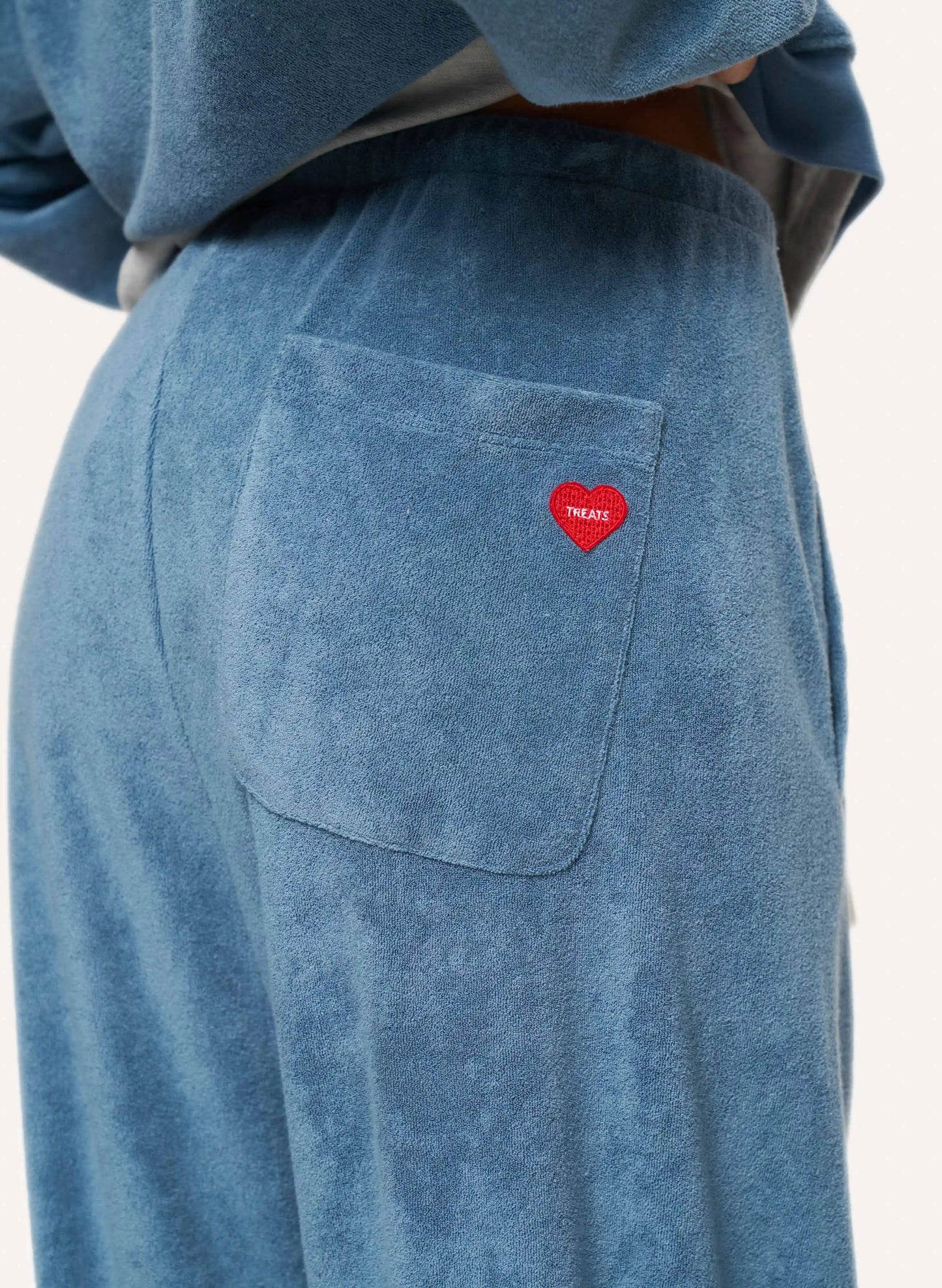 Close-up of a person wearing dusty blue textured lounge pants with a side pocket featuring a small red heart patch that reads 'TREATS'.