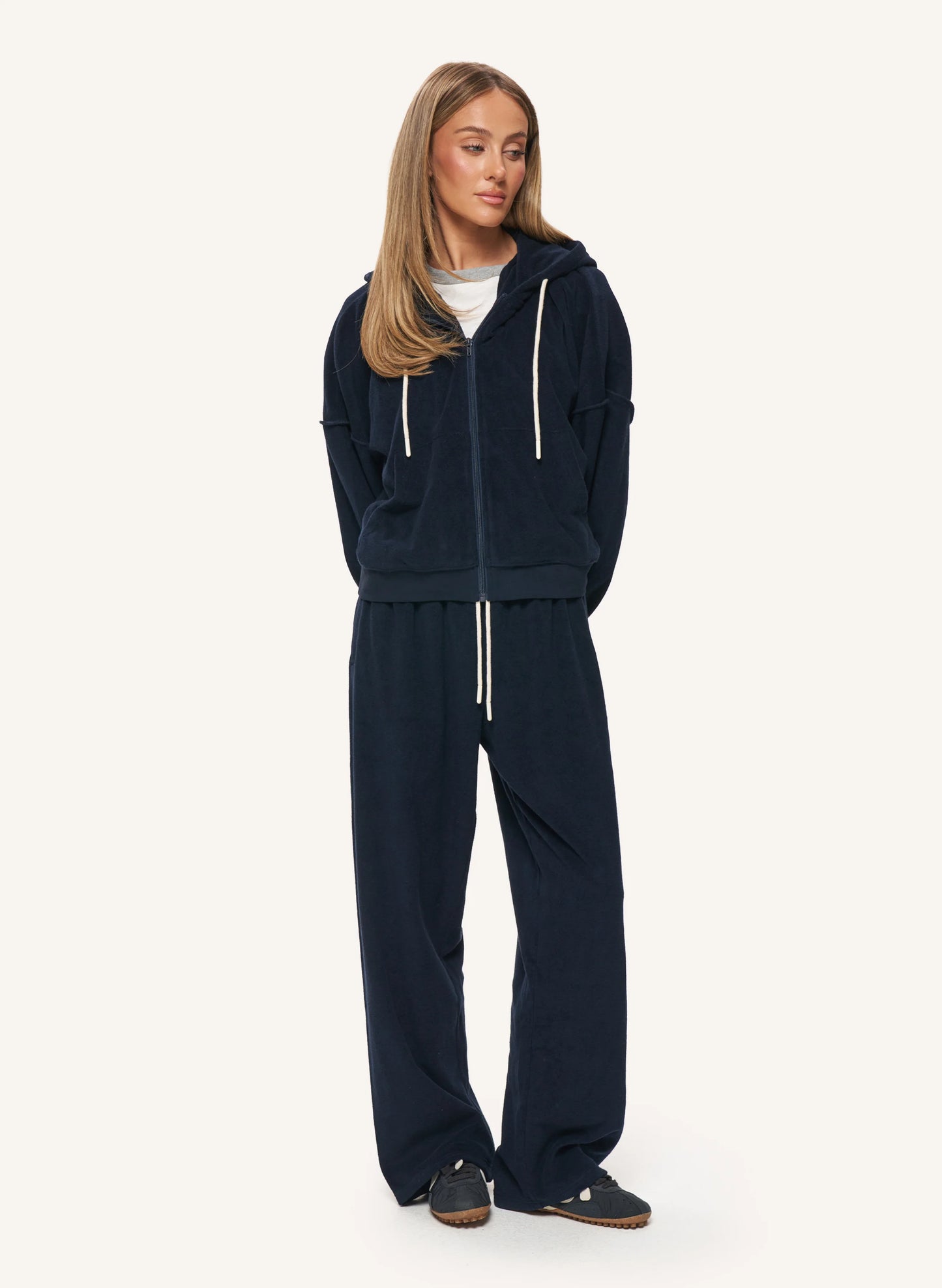 A woman modeling a matching navy blue velour lounge set, consisting of a zip-up hoodie with white drawstrings and matching wide-leg pants, against a plain off-white background.
