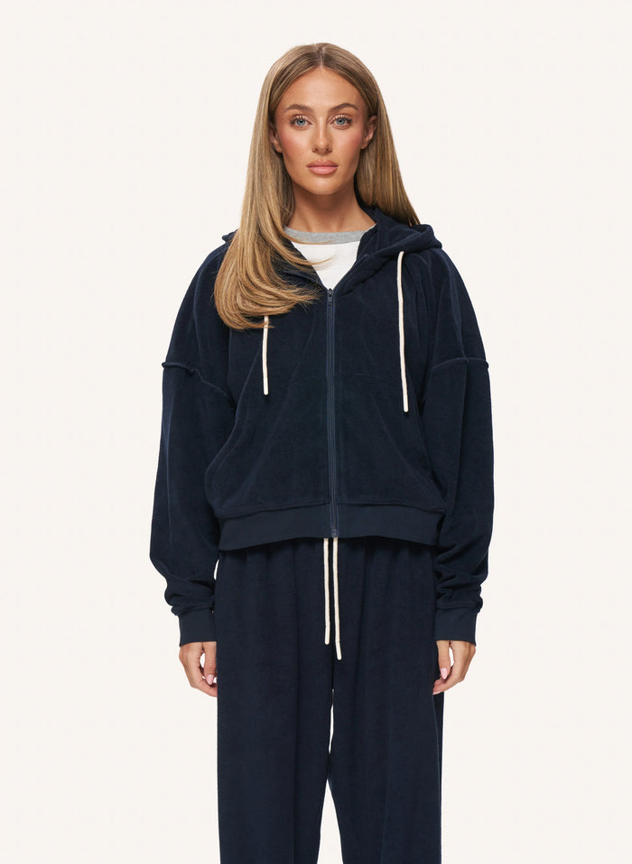A woman modeling a dark navy blue zip-up hoodie and matching sweatpants set with white drawstrings, photographed against a clean off-white studio background.