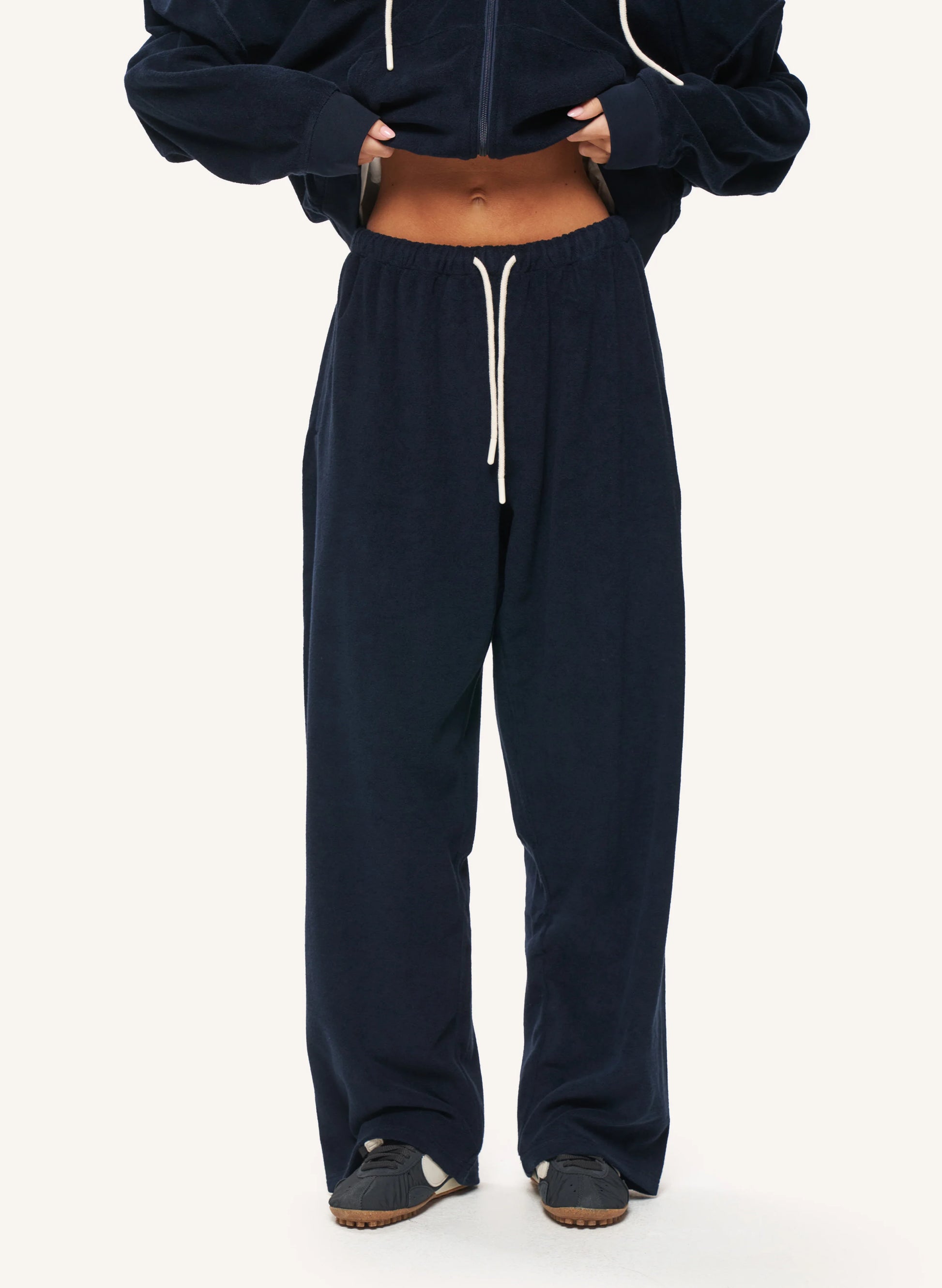 A person wearing loose navy jogger sweatpants with a white drawstring, paired with a cropped navy zip-up hoodie and dark slip-on shoes, photographed against a plain light background.