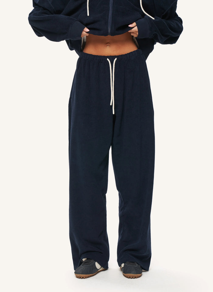 A person wearing loose navy jogger sweatpants with a white drawstring, paired with a cropped navy zip-up hoodie and dark slip-on shoes, photographed against a plain light background.