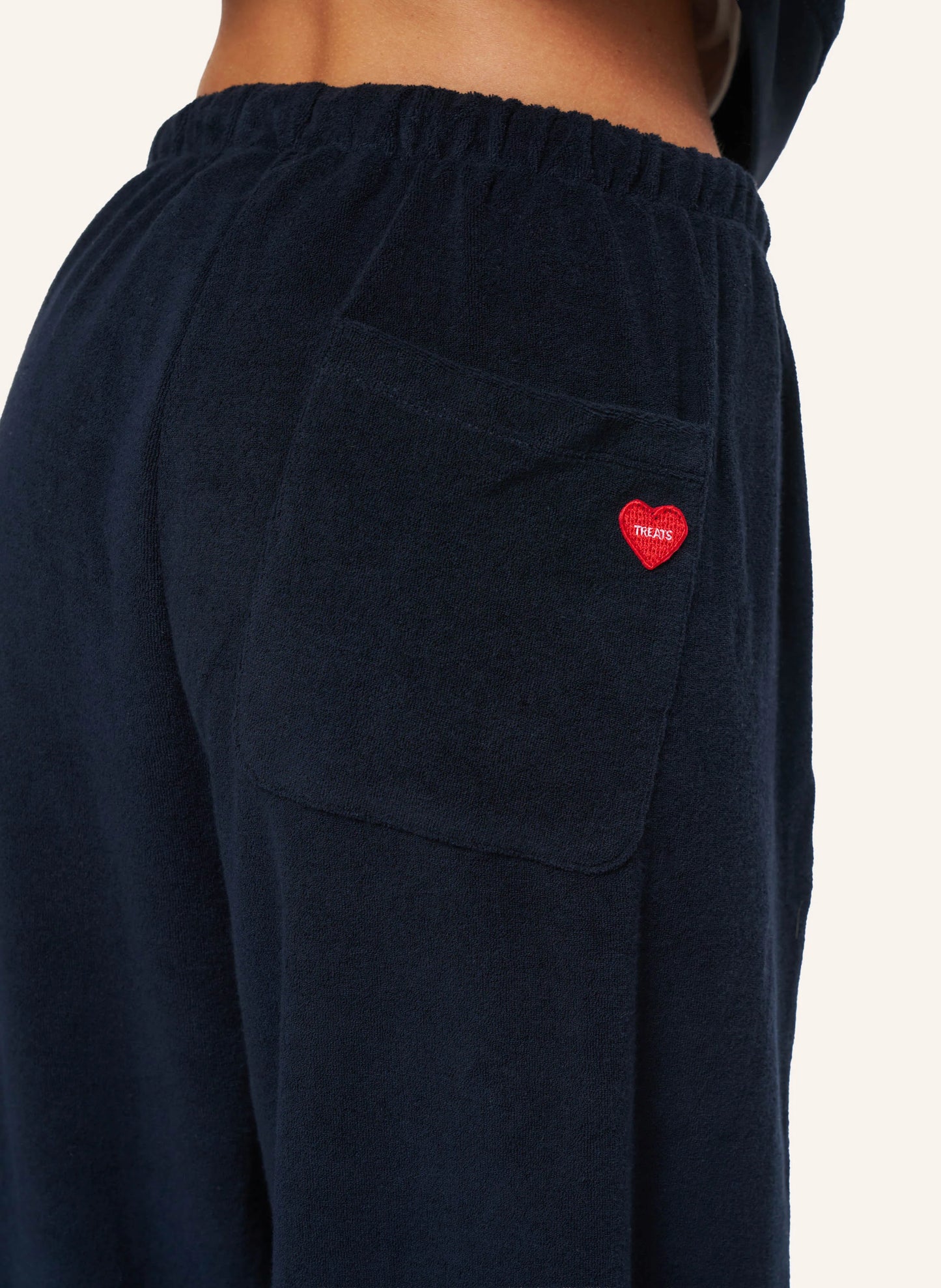 Close-up of a person wearing dark navy lounge pants with an elastic waistband, a side pocket, and a small red heart patch on the pocket. The brand name 'THARSIS' is visible on the heart patch.