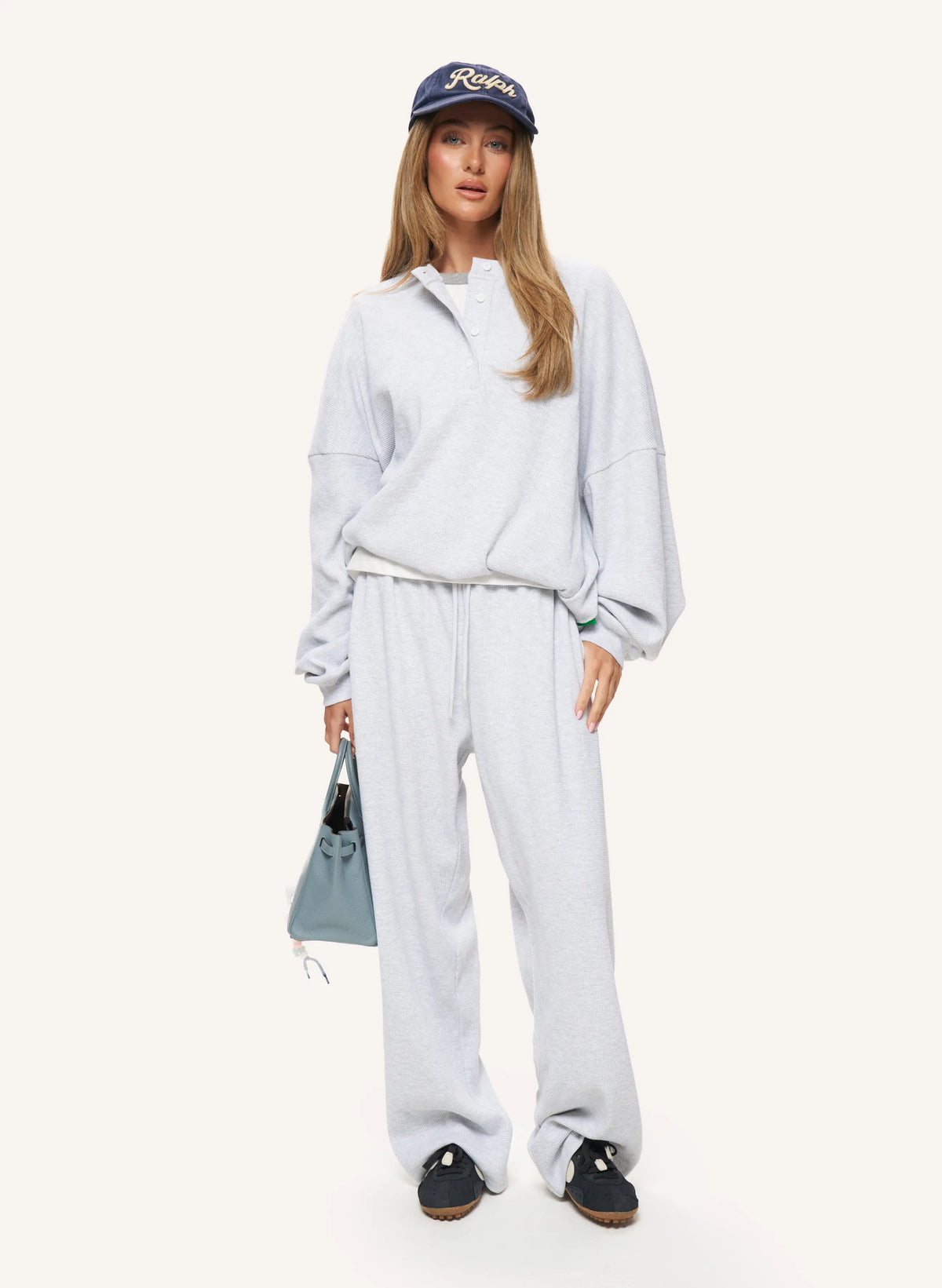 A woman modeling a light gray heathered lounge set consisting of a half-zip sweatshirt and matching sweatpants, paired with a navy blue baseball cap, black slip-on shoes, and a light blue handbag, against a solid off-white background.