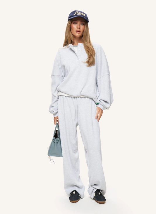 A woman modeling a light gray heathered lounge set consisting of a half-zip sweatshirt and matching sweatpants, paired with a navy blue baseball cap, black slip-on shoes, and a light blue handbag, against a solid off-white background.