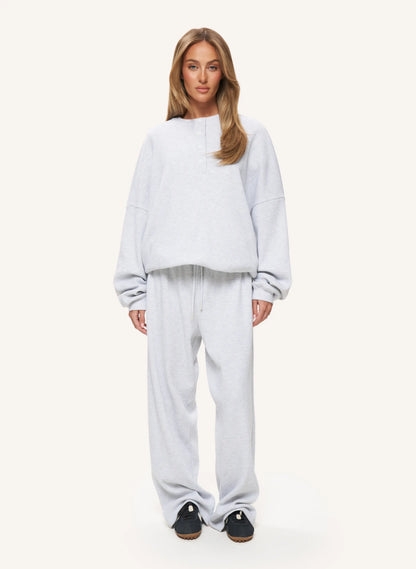 A woman modeling a light gray heathered lounge set consisting of a cropped long-sleeve sweatshirt and matching sweatpants, paired with black slip-on shoes, against a solid off-white background.