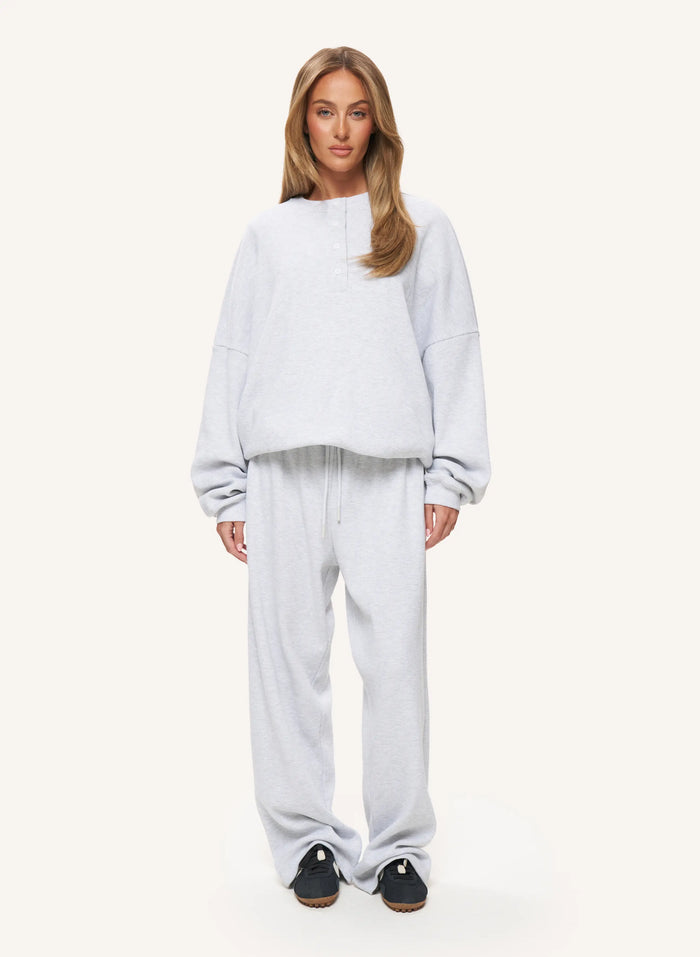 A woman modeling a light gray heathered lounge set consisting of a cropped long-sleeve sweatshirt and matching sweatpants, paired with black slip-on shoes, against a solid off-white background.