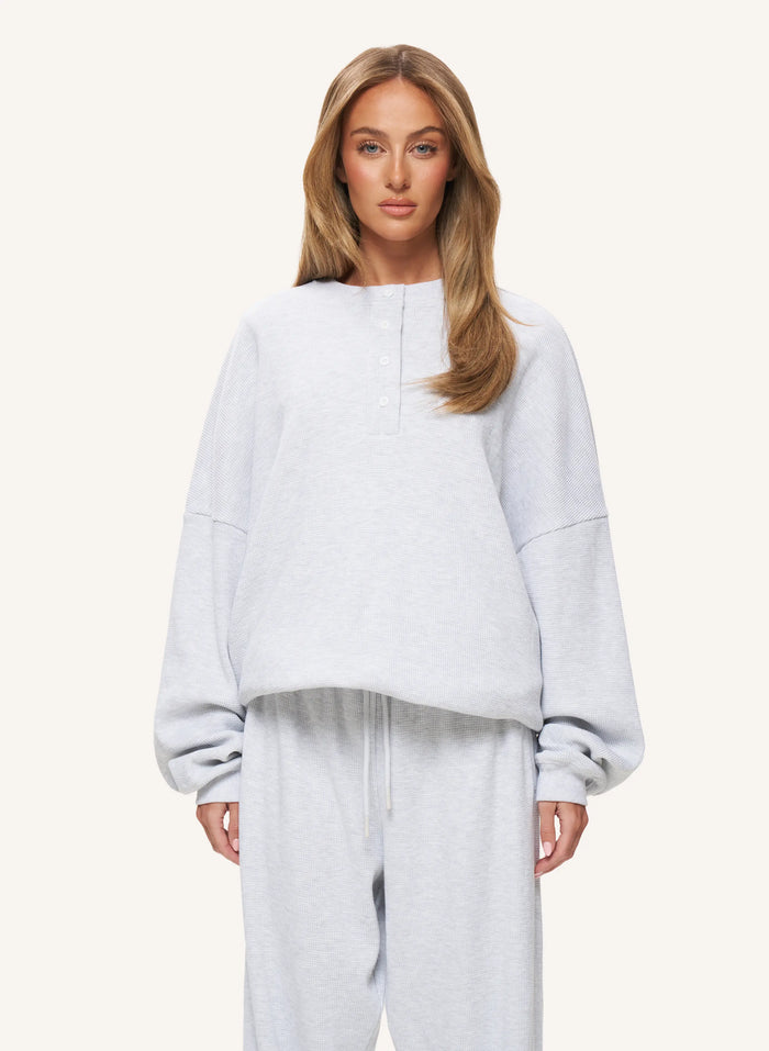 A woman modeling a light gray heathered lounge set consisting of a cropped long-sleeve henley sweatshirt and matching sweatpants, set against a solid off-white background.