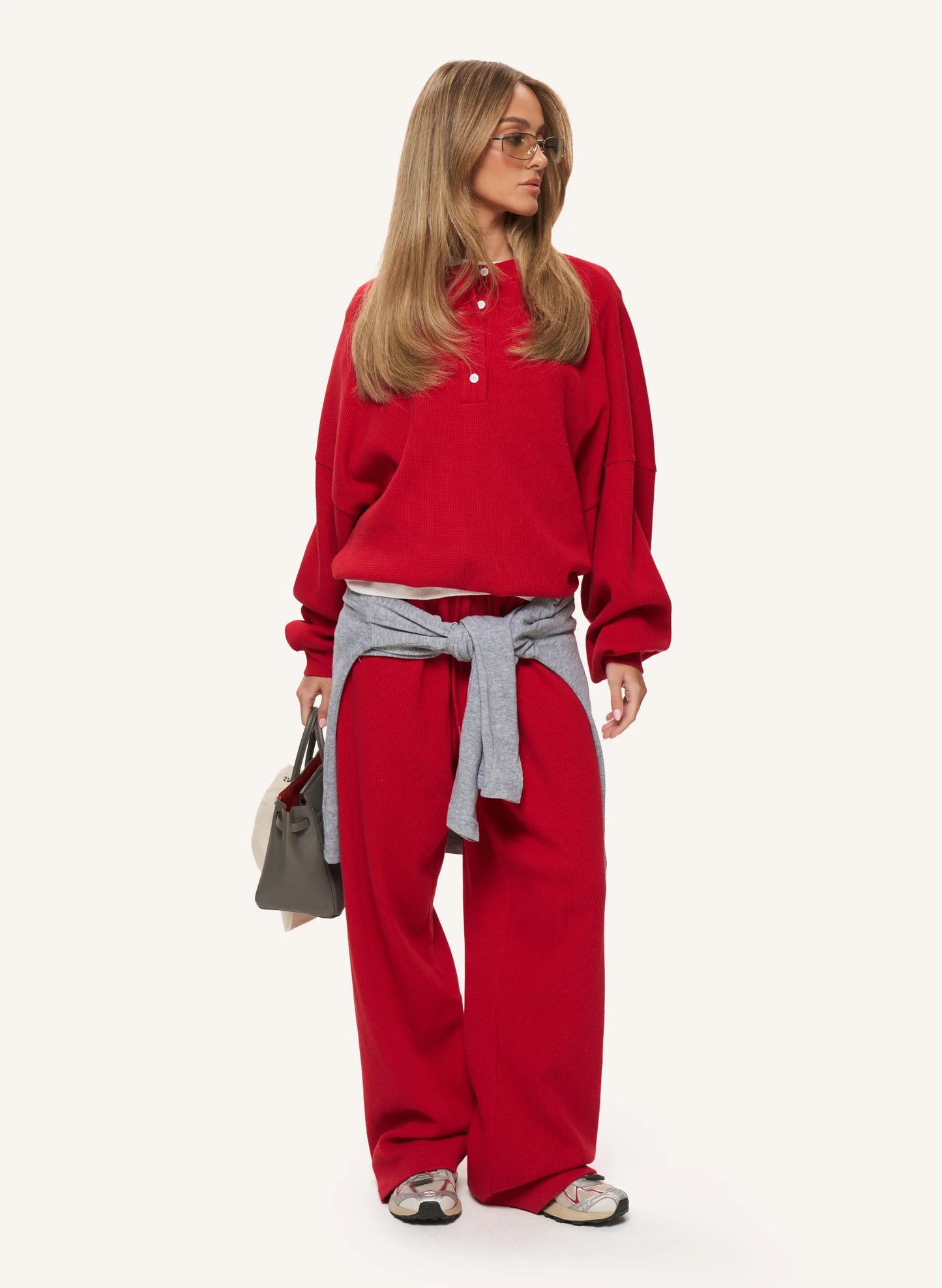 A woman modeling a red long-sleeve polo sweatshirt and matching red wide-leg sweatpants, with a gray tie-waist sash, glasses, and a small gray handbag against a plain light background.