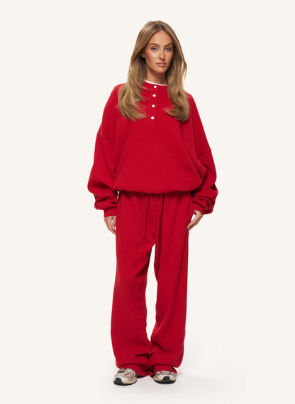 A woman modeling a matching red lounge set consisting of a cropped henley sweatshirt with white buttons and matching wide-leg sweatpants, set against a plain off-white studio background.
