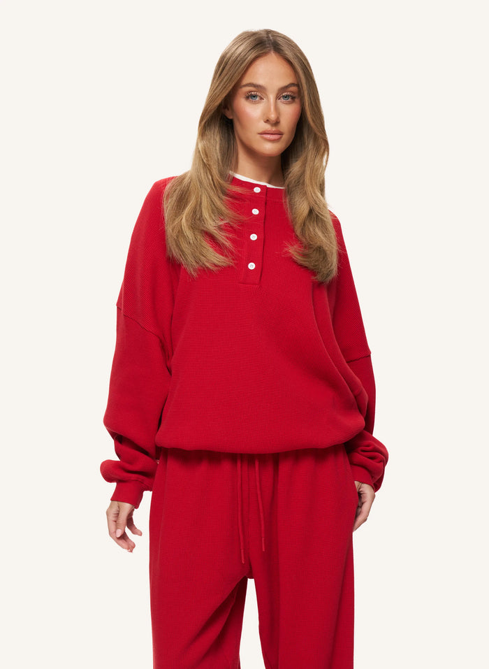 A woman modeling a bright red two-piece loungewear set, featuring a cropped henley sweatshirt with white buttons and matching drawstring sweatpants, set against a plain off-white studio background.