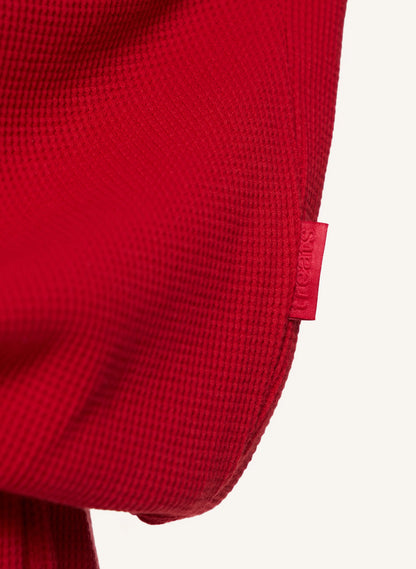 Close-up of a bright red textured garment, showing a small red fabric brand tab on the side against an off-white background.