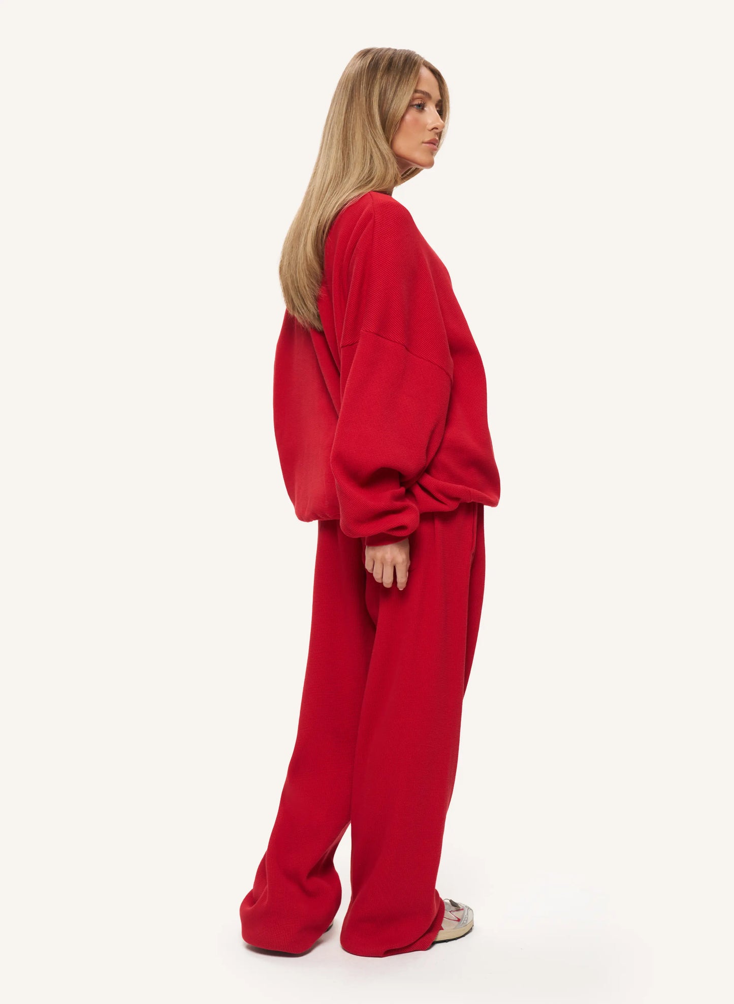 A woman wearing a matching red lounge set consisting of a loose long-sleeve sweatshirt and wide-leg trousers, shown from a side-back angle against a plain off-white studio background.