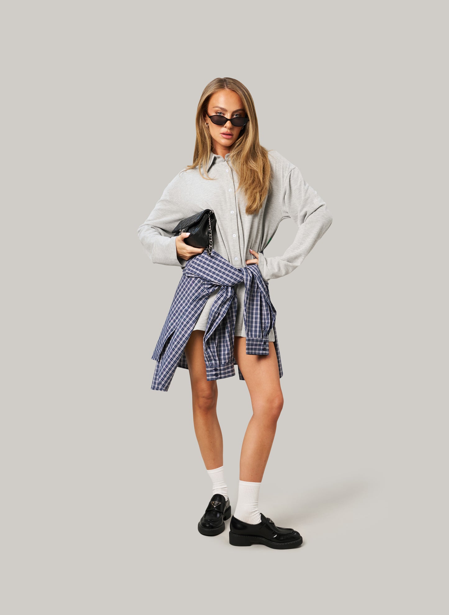 Woman wearing a light grey romper, plaid skirt, and black shoes on a plain background